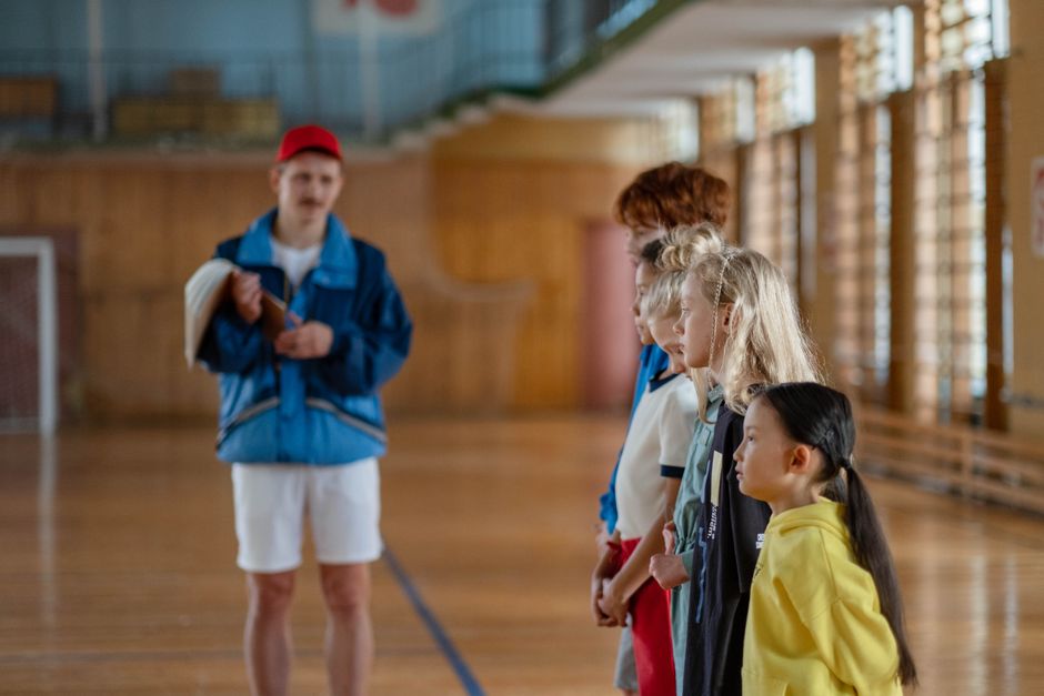 Diverse group of children standing indoors in a gym with a coach in focus.