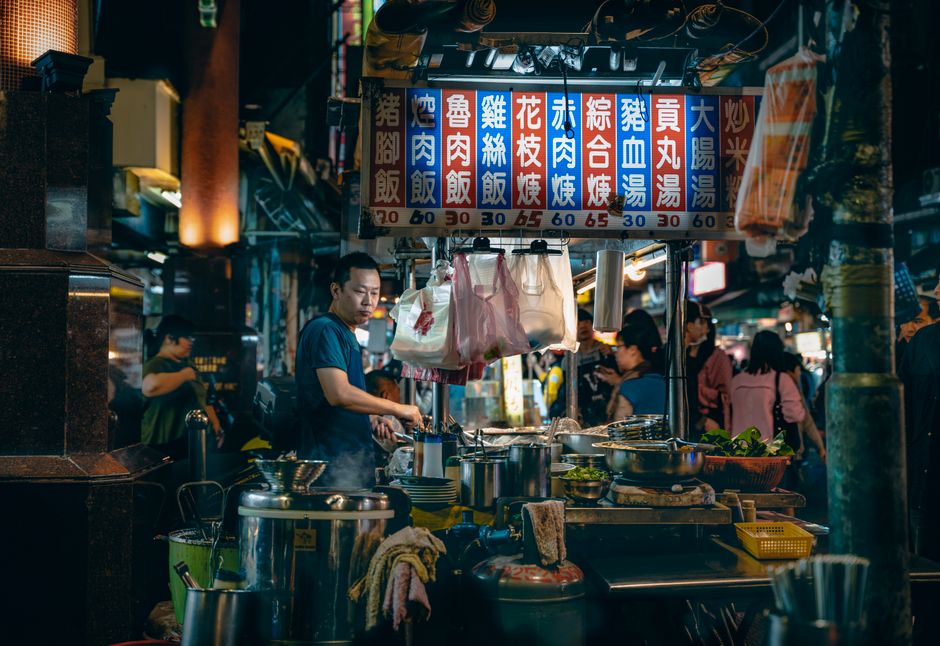 Side view of thoughtful male cook looking down while preparing food at table with bowls under colorful signboard with hieroglyphic inscriptions and numbers near customers at night