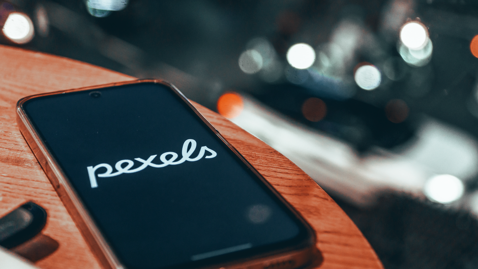 Smartphone on wooden table displaying Pexels logo, blurred bokeh lights in background.