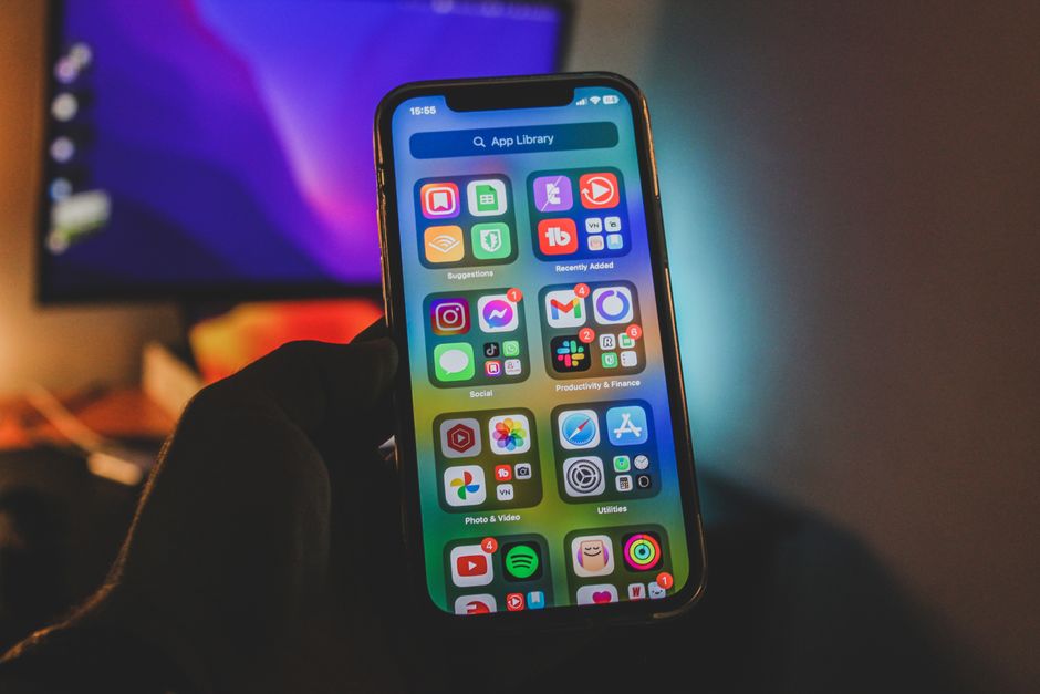 A hand holds a smartphone showing a colorful app-filled screen in an indoor room with dim lighting.